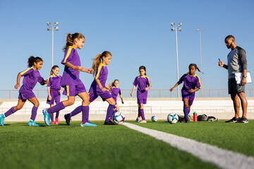 Girls in soccer uniforms on field with coaches, each with a ball, during training session.