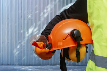 Worker holding orange safety helmet with protective earmuffs for construction and industry. Industrial safety, construction equipment, and protective workwear against noise and hazards.
