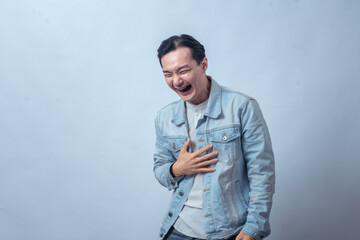 Asian man in denim jacket laughing hard, holding his chest with hand while closing eyes, showing...