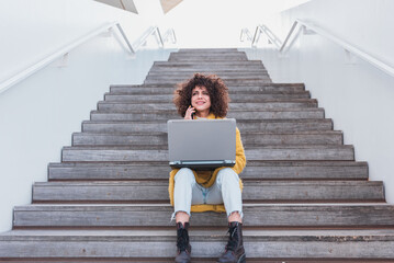 Smiling Latin woman freelance working remotely using laptop and smartphone while sitting on stairs