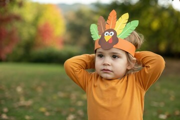 Child wearing handmade turkey headband costume for Thanksgiving celebration in autumn park with copy space.