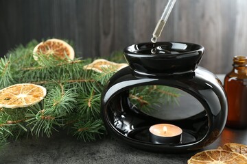 Dripping essential oil from pipette into aroma lamp with burning candle at grey textured table against wooden background, closeup