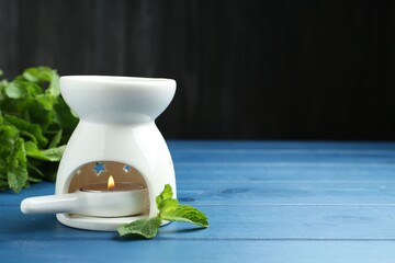Aroma lamp with burning candle and mint leaves on light blue wooden table against dark background, closeup. Space for text