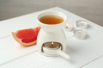 Aroma lamp with burning candle and slice of grapefruit on white wooden table against grey background, closeup