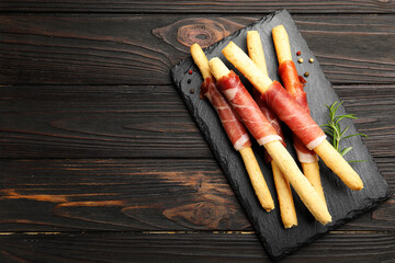 Delicious grissini sticks with prosciutto, peppercorns and rosemary on wooden table, top view. Space for text