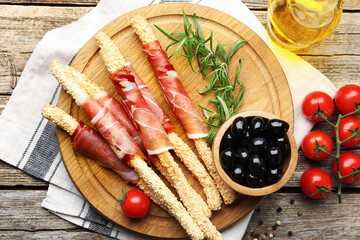 Delicious grissini sticks with sesame seeds, prosciutto and other products on wooden table, flat lay