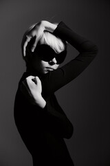 Obraz premium Stylish woman in black outfit striking a pose with bold sunglasses, showcasing confidence and fashion in a dramatic black and white setting.