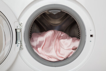 Pink silk fabric in opened white washing machine. Delicate clothes care. Closeup. Front view.