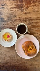 flat lay of cozy breakfast scene featuring a cup of black coffee, a chocolate pastry over a rustic wooden table. Warm natural lighting highlights the textures and creates a comforting atmosphere.