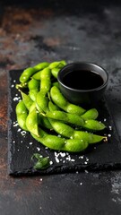 Roasted edamame pods with a dark soy dipping sauce on a black slate tray