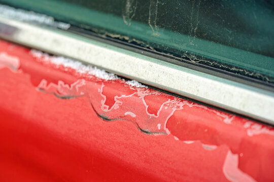 Damaged red car paint on door with peeling clear coat and faded paintwork, oxidation, deterioration, and visible surface wear on the exterior finish of old vehicle. Selective focus