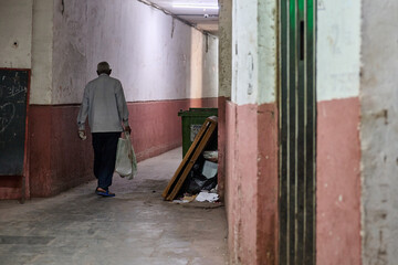 A narrow hallway with dim lighting in Dharavi, Mumbai, showing an elderly man walking down with a bag in hand, with scattered trash and a worn-out environment. T