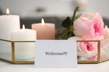 Card with word Welcome, roses and burning candles on white table, closeup