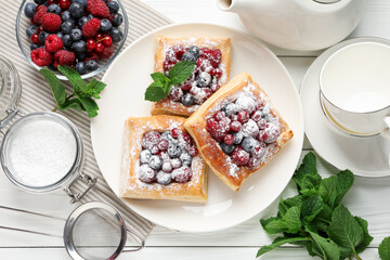 Tasty puff pastries with berries, powdered sugar and mint on white wooden table, flat lay