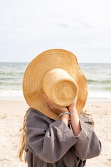 young woman on the beach holding a straw hat infront of her face