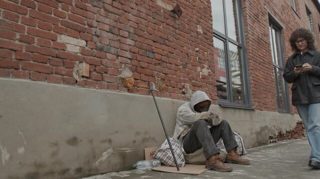 African American homeless man sitting near brick wall with his head down, he holding metal cup while begging for money in city street, people walking past him
