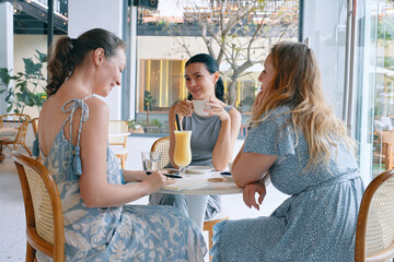 Group of young female friends is sitting at table in a cafe with variety of drinks and talking with happy smiles.