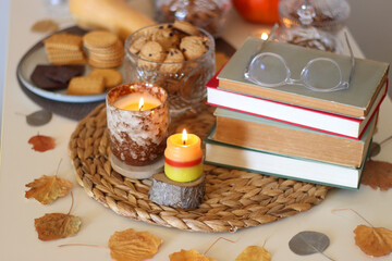 Lit candles, cookies, chocolate, nuts, tea, wine, pumpkins, books, reading glasses and autumn leaves on the table. Autumnal hygge at home. Selective focus.