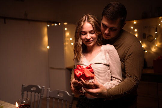Concept of Happy Valentine's Day. Young couple in love celebrating anniversary at home. Husband gives gift box to his wife.