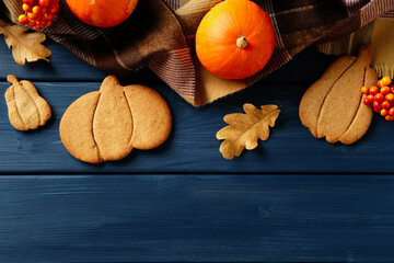 Fototapeta premium Festive autumn flat lay with pumpkin cookies, plaid blanket, pumpkins, berries, and oak leaves on dark blue wood table.