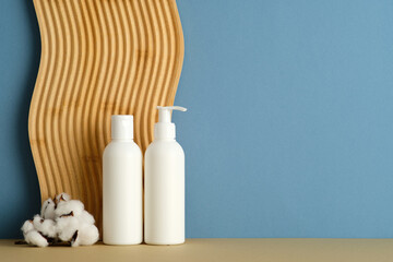 Two white bottles with pump and flip cap on beige surface, cotton bolls, wavy wooden panel, blue background, clean skincare concept.