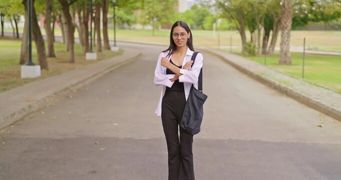 Indian happy young adult gen z proud cute woman look camera cross arms standing campus area. Teen pupil smart girl wear glass bag fold hand staring cam do casual pose enjoy new day garden place
