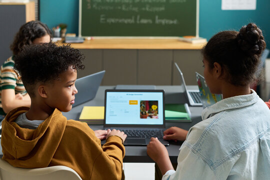 Black boy and Black girl sitting at desk using laptop computers in classroom, engaging in collaborative learning activity with other students visible in background