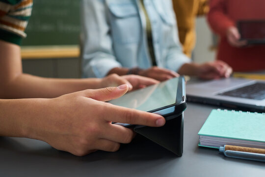 Teenager hands holding tablet device on desk in classroom setting, with multiethnic group of teenagers standing in background, notebook and laptop visible on table