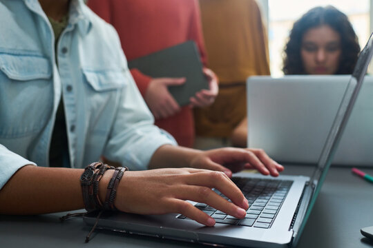 Young adult Caucasian woman typing on laptop with multiethnic group of teenagers working together in background, focusing on collaborative project or study session in modern setting - Powered by Adobe