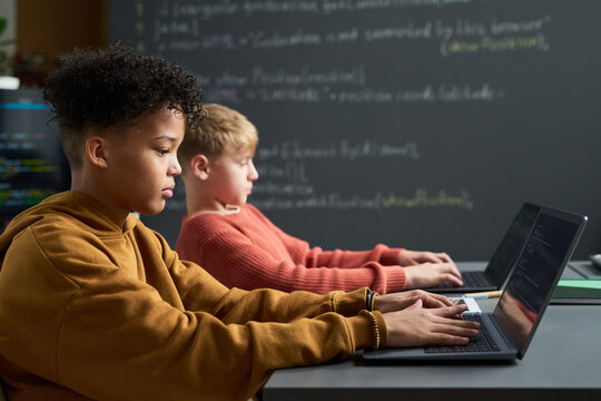 Multiethnic boys sitting side by side using laptops in classroom, focusing on typing and coding, with chalkboard and programming code visible in background, representing technology education