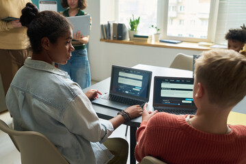 Teenage Black girl and teenage Caucasian boy sitting at table using laptops, collaborating on project in modern classroom with other students visible in background