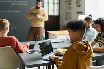 Teenage boys and girls of diverse ethnicities sitting at desks using laptops for coding lesson, while male Caucasian teacher standing in background holding digital tablet in classroom