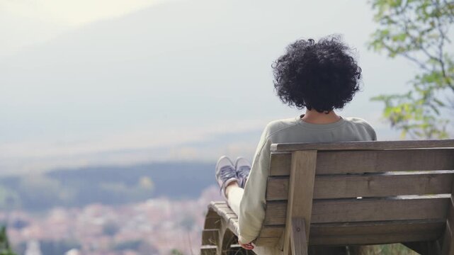 A woman sits on a bench overlooking a mountain and aerial cityscape view of the city. The scene is peaceful and serene, with the woman enjoying the view and the quiet atmosphere
