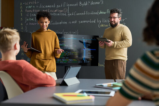 Teenager boy presenting programming project using tablet while standing near Caucasian male teacher holding digital tablet in classroom with students listening