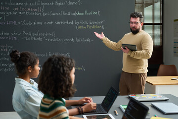 Caucasian man explaining programming code on chalkboard while holding tablet, two teenage girls and...