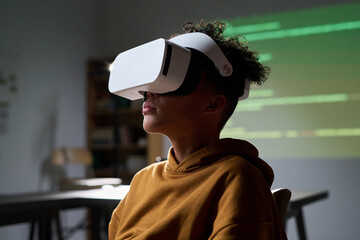 Teenager Black boy wearing virtual reality headset sitting indoors, experiencing immersive digital...