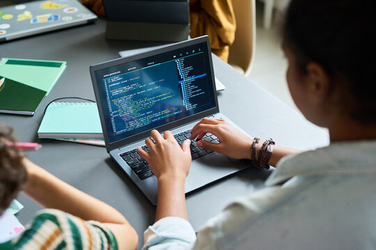 Teenage Black girl coding on laptop, sitting at desk with hands on keyboard, focusing on programming task, other student partially visible nearby, educational setting