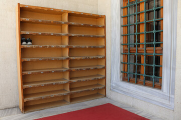 Wooden shoe rack with numbered shelves near a window and red carpet at a mosque entrance. Fits topics of etiquette, cleanliness, organization, worship space and culture.