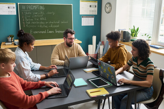 Group of diverse teenagers collaborating with Caucasian man using laptops in classroom, engaging in technology-based learning, sitting around table with notebooks and digital devices