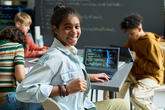 Portrait of Black teenage girl smiling at camera while sitting at desk with open laptop displaying code, multiethnic teenagers working together on computers in classroom background