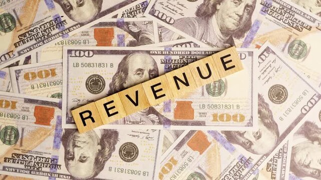 Wooden blocks with the letters "Revenue" arranged on a slowly rotating stack of dollar bills. Looping 360-degree of wooden blocks with the words "Revenue" as a business financial concept. 4K footage.