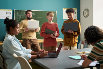 Diverse group of teenagers and young adults collaborating in classroom, Caucasian man and Black teenager standing with laptops, two young women working at table, chalkboard in background
