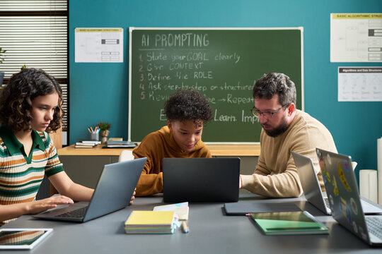 Caucasian man assisting Black teenager and Caucasian teenage girl working on laptops in classroom setting, chalkboard with AI prompting instructions visible in background - Powered by Adobe