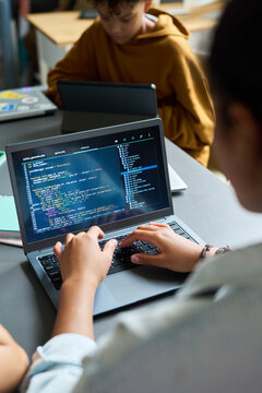 Teenager typing code on laptop, sitting at desk in classroom, another teenager with curly hair using laptop in background, focusing on programming activity