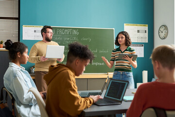 Multiethnic group of teenagers sitting at desks using laptops while young adult Caucasian man and young adult woman standing near chalkboard leading classroom discussion