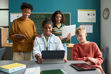 Group of multiethnic teenagers collaborating on digital tablets and laptop in classroom, smiling and interacting with technology, diverse students engaging in educational activity together