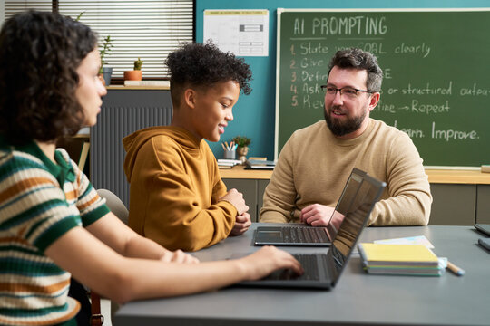 Caucasian man teaching multiethnic teenagers using laptops in classroom, students listening and engaging in discussion, chalkboard with AI prompting instructions visible in background - Powered by Adobe