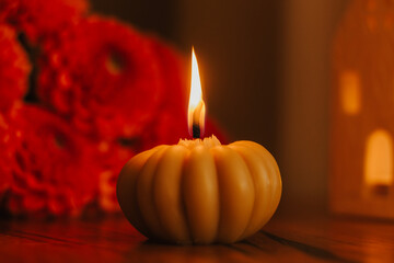Pumpkin-shaped candle with flame and orange chrysanthemum flowers on wooden table. Warm autumn still life symbolizing cozy home decor, festive mood, relaxation, and seasonal atmosphere.
