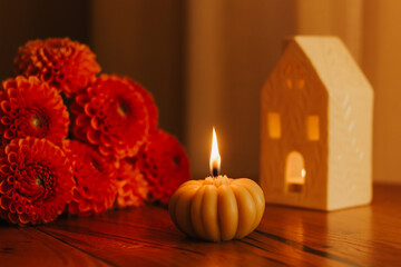 Autumn still life with orange dahlia flowers, burning pumpkin candle and ceramic house lantern on wooden table. Cozy home decor, fall vibes, hygge atmosphere, Thanksgiving or Halloween concept.