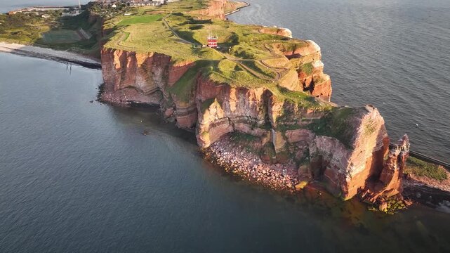 Aerial footage of island Helgoland in North Sea, Germany with striking red sandstone cliffs, rock pillar &ldquo;Lange Anna&rdquo; rises from water next to the island, surrounded by seabirds and rugged coastline.
